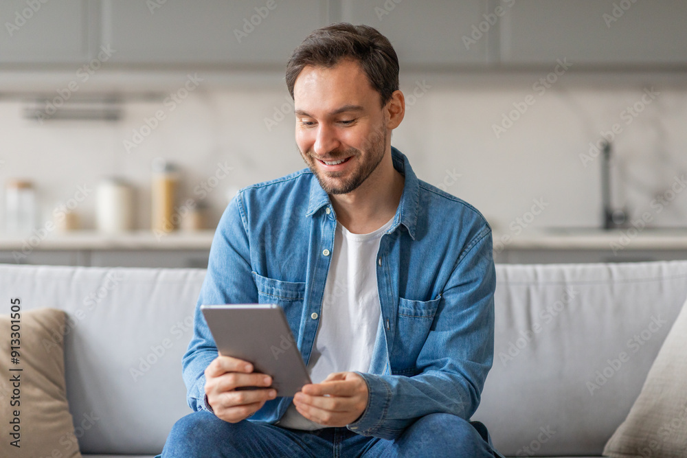 Poster a man in a blue shirt and jeans sits on a light gray couch in a living room. he is looking down at a