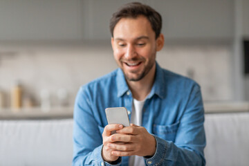A man sits in a kitchen, wearing a blue button-up shirt and white undershirt, looking down at his smartphone with a wide grin. He is excited by what he is reading.