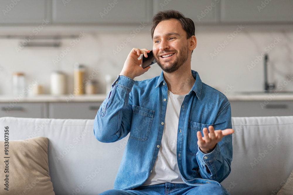 Poster a man with a beard is sitting on a white couch in his home while talking on his phone. he is wearing