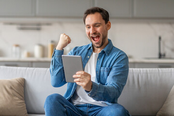 A man sits on a white couch in a living room and celebrates with a raised fist while looking at a tablet in his hands. He is wearing a white t-shirt and a blue denim shirt.