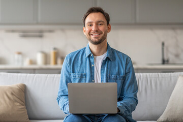 A man with short brown hair and a beard sits on a white couch, wearing a blue denim shirt and jeans. He is smiling and looking at the camera, using a laptop computer. .