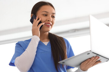 Young female medical receptionist holding modern laptop and talking on mobile phone in hospital