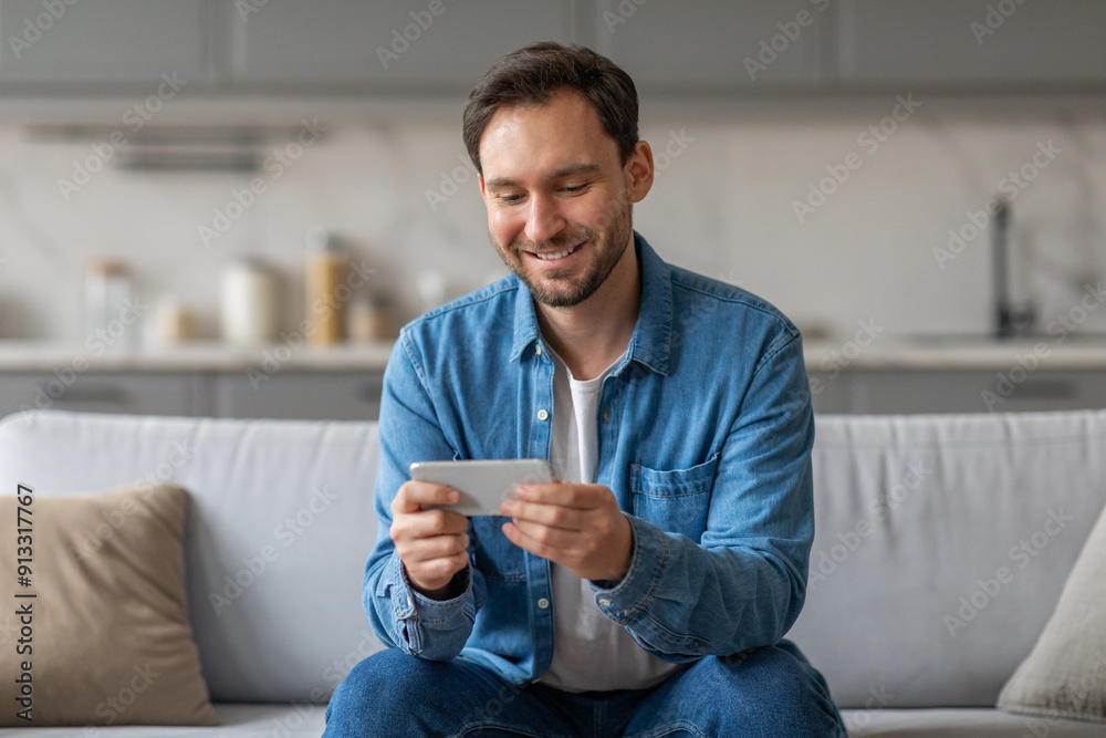 Poster A man with a light beard sits on a white couch in a modern home. He is smiling as he uses his smartphone. The kitchen is visible in the background.