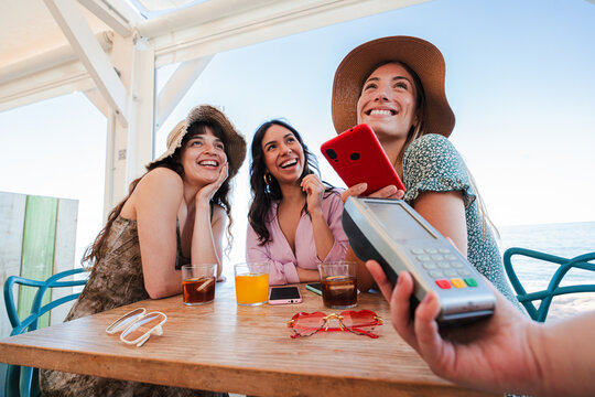 Happy young group of only women paying the bill with a nfc cellphone in cocktail beach bar. Female holding a smart phone and giving a payment transaction to the cashier on vacations with her friends