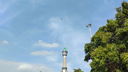 A low-angle view of the mosque tower next to a banyan tree and city lampposts against a blue sky with wispy clouds.