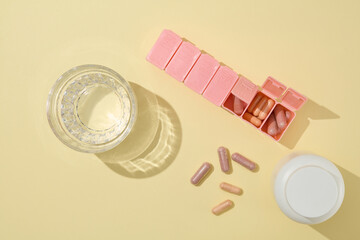 Idea to design pharmaceutical advertisement poster, a pink pill box contains vitamin capsules displayed on yellow table top, next to white medicine jar and a glass of water. Top view, space for text