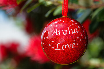 A shiny red Christmas bauble with the words aroha and love, hanging in a Summer flowering New Zealand Pohutukawa tree. The background is defocused.