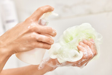 female hands applying liquid soap shower gel from white cosmetic bottle on sponge in bathroom