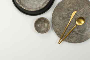 Top view of brown marble tray, plate and bowl, black plate, gold cutlery on white background. Food presentation, dishes, stone tray. Copy space, flat lay.