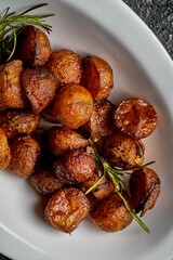 A close-up of roasted baby potatoes in a white oval dish. The potatoes are golden brown and crispy, garnished with fresh rosemary.
