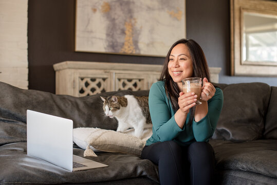Happy asian woman sitting on couch with laptop and cat