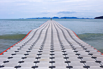 Long, floating walkway stretches over calm sea waters towards distant islands under cloudy sky,...
