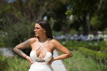 A young, dark-haired, beautiful woman, heavily tattooed, in a white dress and sunglasses with purple crystals, sitting, with her hands on her waist, looking into infinity relaxed and calm.