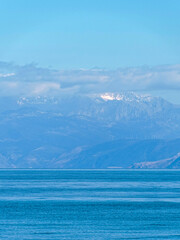 Blue sky and sea with some clouds above the snowy mountains in between. Slight view of some wind generators on the slopes.