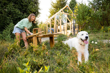 Female carpenter standing by a workbench and her large white dog in front of a partially built wooden greenhouse in her backyard. Engaged in a DIY project, surrounded by nature