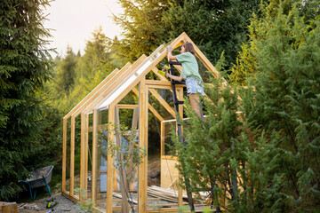 Female carpenter using a power drill while working on a wooden greenhouse in her backyard, engaged in a DIY project, surrounded by nature on sunset