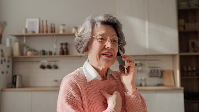 Modern older Caucasian grandmother holding smartphone while having phone call. Pretty elderly woman friendly communicating from smartphone in cosy kitchen at home. Calling friends or relatives.