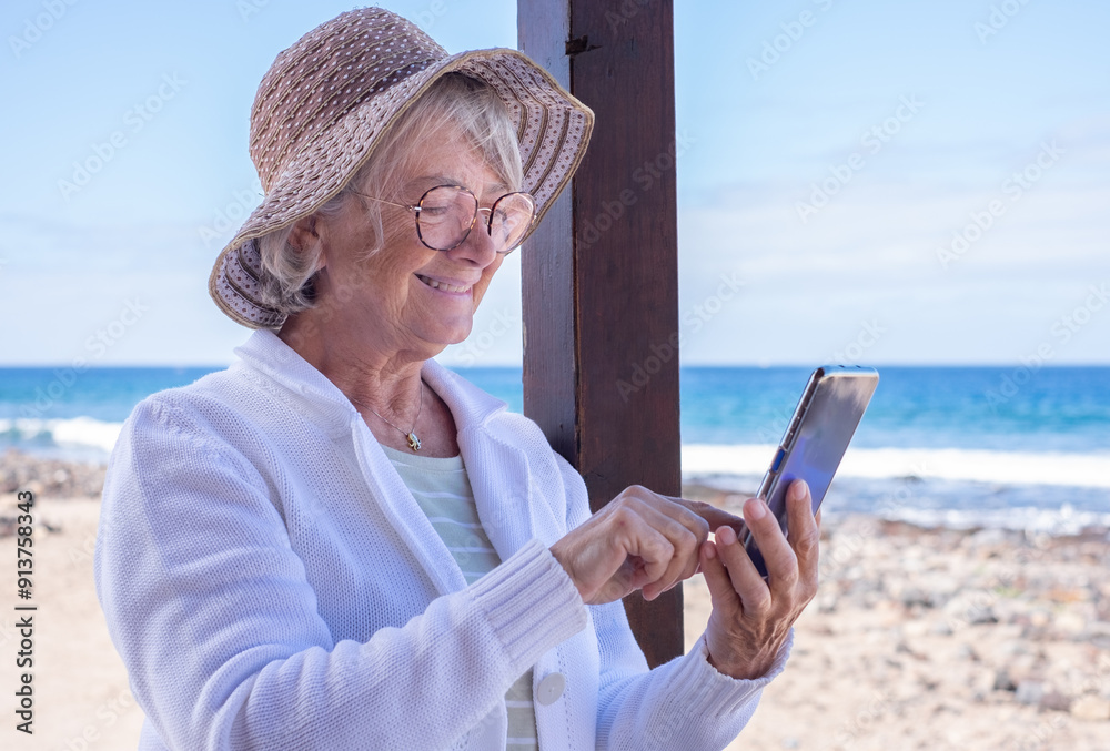 Canvas Prints smiling senior woman wearing hat and sunglasses standing at the beach using mobile phone typing a me