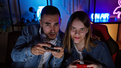 A man and woman are engaged in gaming together in a dark room illuminated by neon lights at home.