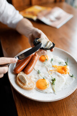 handsome man is eating a delicious English breakfast with juicy sausages, fresh vegetables, fragrant herbs, and scrambled eggs on a beautiful white plate in a cozy nice restaurant