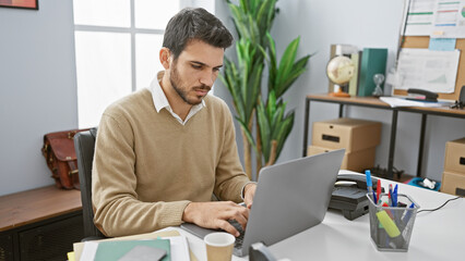 A young hispanic man with a beard works on a laptop in a modern office setting, portraying professionalism and focus.