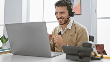Handsome hispanic man with beard using laptop and headset in a bright office setting, exuding friendly professionalism.