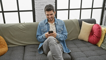A young hispanic man with a beard smiles while using a smartphone on the sofa in a cozy living room.