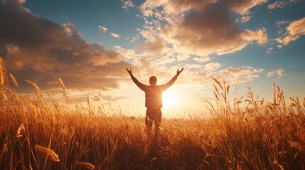 A man stands in a golden field, arms raised high in prayer, basking in the warm glow of a stunning sunset with clouds painted in vibrant colors