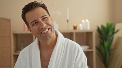 Middle-aged hispanic man relaxing at an indoor spa and wellness center, looking content in a white bathrobe surrounded by candles and soft lighting.