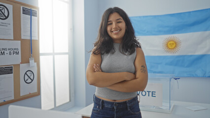 Young woman with crossed arms smiling in an indoor voting room with an argentine flag and voting...