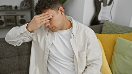 Stressed young man with curly hair sitting on a couch, covering his face with his hand in a modern living room.