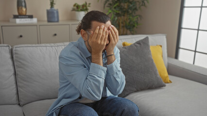 Hispanic man with moustache sitting on couch in living room, hands covering face, conveying emotions in a modern home interior.