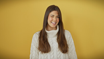 A cheerful young hispanic woman in a white sweater poses against an isolated yellow background, exuding joy with a playful wink.