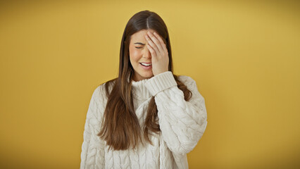 A young hispanic woman in a white sweater poses with a smile, touching her head against an isolated yellow background.