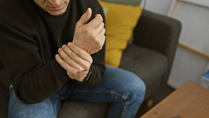 Man suffering from wrist pain while sitting on a couch at home, indicating discomfort and health...