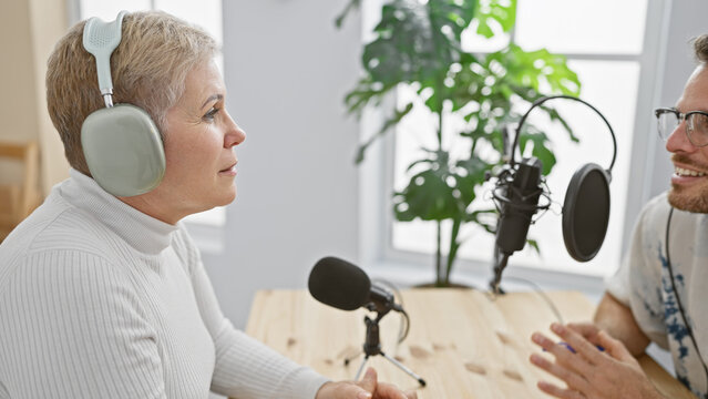 A woman and a man converse indoors during a podcast recording session surrounded by equipment.