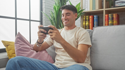 A smiling young man using a smartphone while comfortably seated on a sofa in a well-lit home interior.