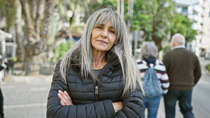 Mature hispanic woman with grey long hair stands arms crossed on a busy urban street.