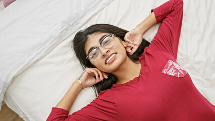 Naklejka premium A beautiful young indian woman with long hair and glasses smiles while lying comfortably on a white bed in a well-lit bedroom.