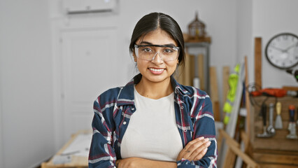 A young indian woman with long hair and safety glasses smiles confidently in a well-equipped carpentry workshop.