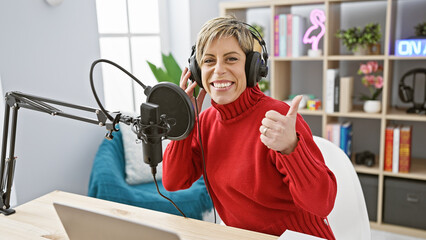 Smiling woman with short blonde hair in a red sweater giving a thumbs-up in a modern radio studio