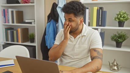 A thoughtful african american man with a tattoo looks worried at home, leaning on a desk with a laptop in a modern room.