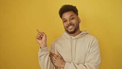 Portrait of a cheerful african american man pointing while wearing a casual hoodie against a yellow...
