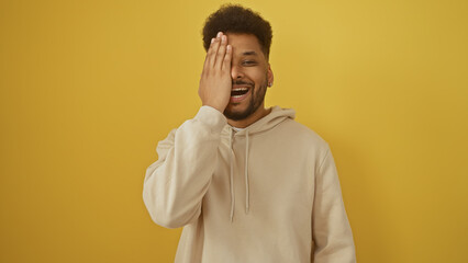 A cheerful african american man in a hoodie smiling with hand on face against a yellow background