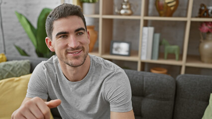 A handsome young man with a welcoming smile sitting in a modern living room, portraying a casual and relaxed home environment.