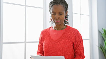 African american woman in office reading document, wearing red, with bright window background.