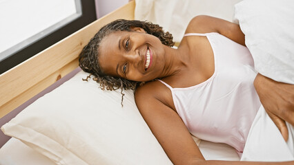 African american woman smiling in bedroom, radiating beauty and comfort while lying in bed, indoors.