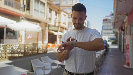 Handsome young hispanic man in a white shirt checking his smartwatch on a street in an urban town setting outdoors.
