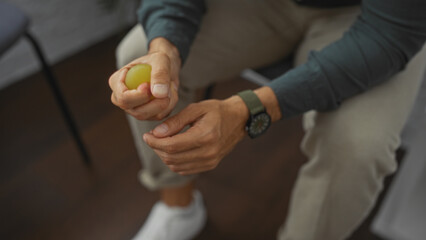 A hispanic man sits in an indoor room, holding a stress ball in his hands, implying an atmosphere of waiting or contemplation.