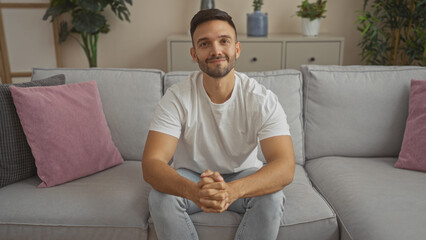 Handsome young hispanic man with a beard sitting in the living room of his home, looking relaxed and cheerful while wearing a white t-shirt and jeans.